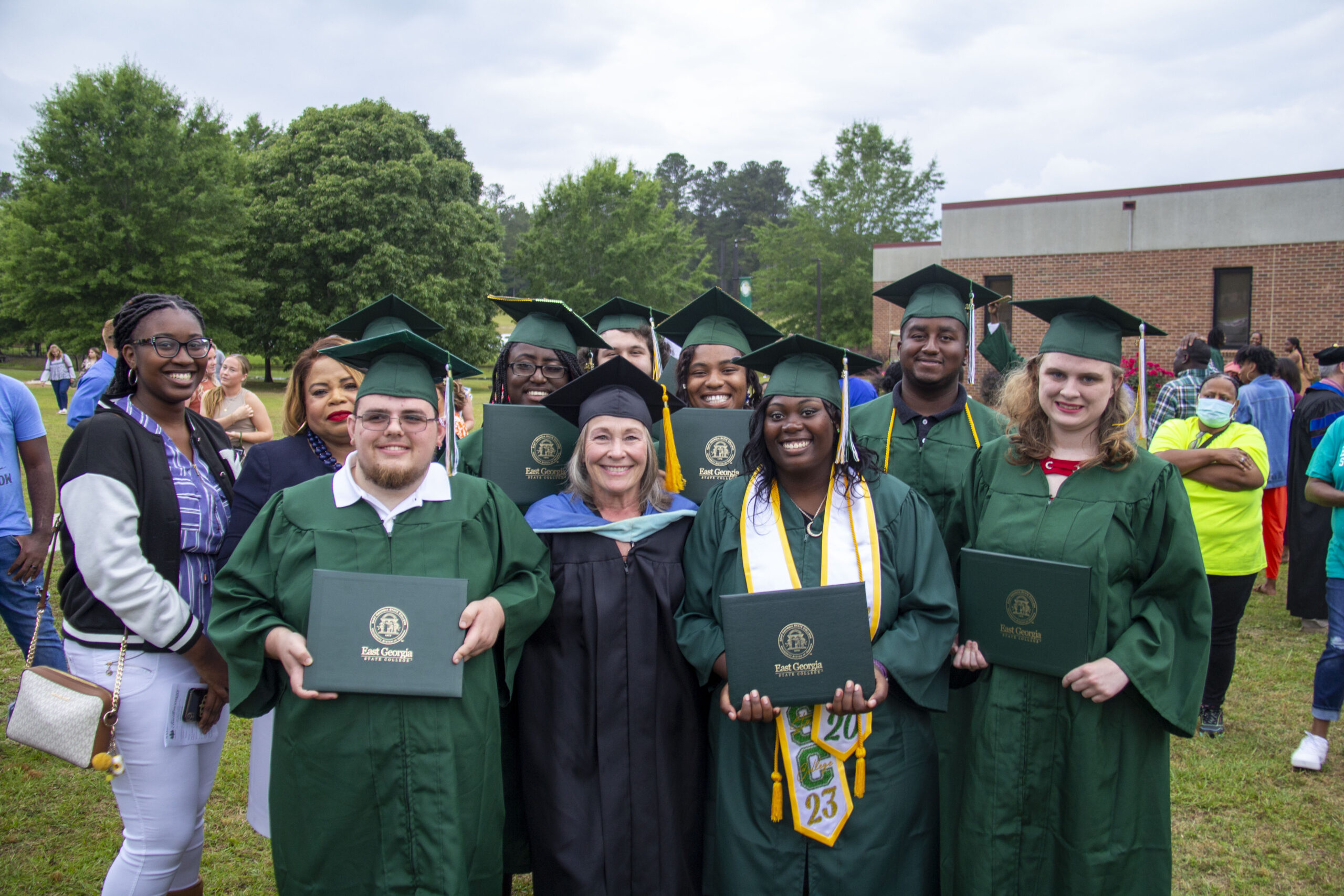 Group of graduates smiling and holding diplomas during an outdoor graduation ceremony.
